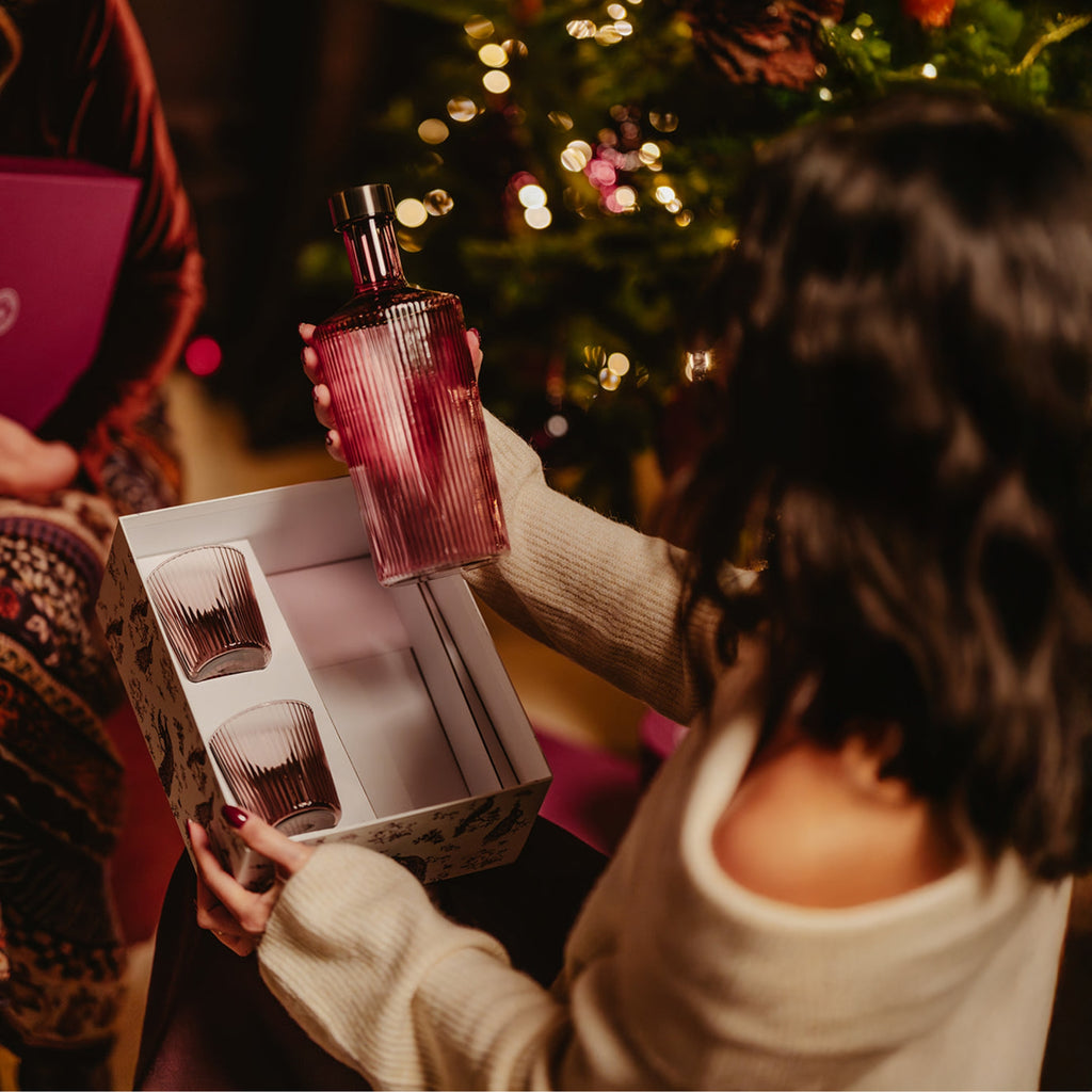 Eine Dame öffnet das Geschenkset Paveau und hält die Paveau Glasflasche in Amethyst in ihrer Hand, neben einem Weihnachtsbaum.